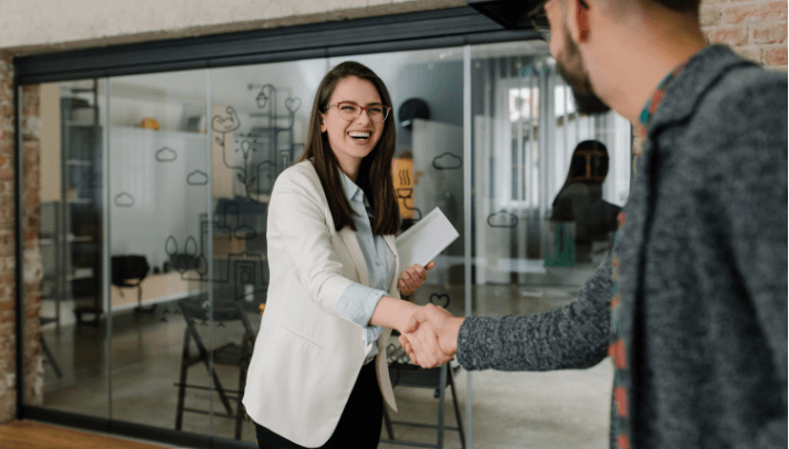 Female professional with dark hair and glasses shakes the hand of a young bearded man wearing a gray jacket