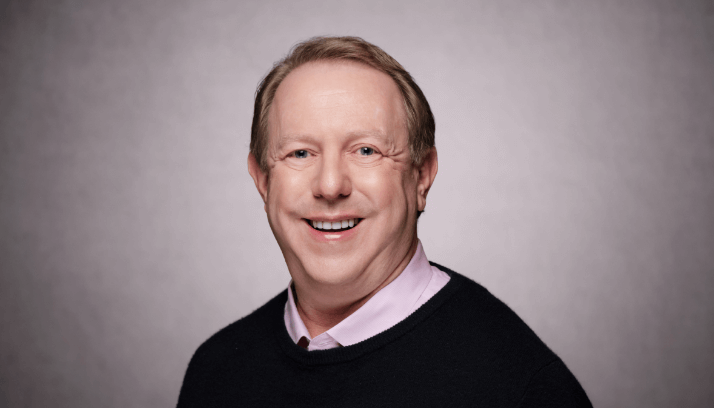 Male business professional in a pale pink shirt under a dark blue sweater against a light brown background
