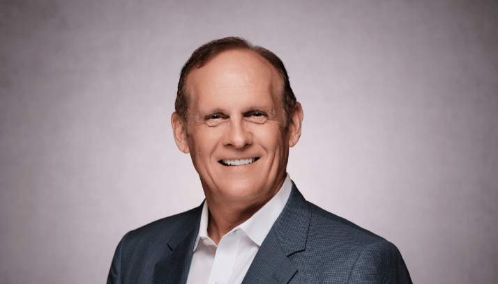 Male business professional in a white shirt and charcoal jacket against a light brown background